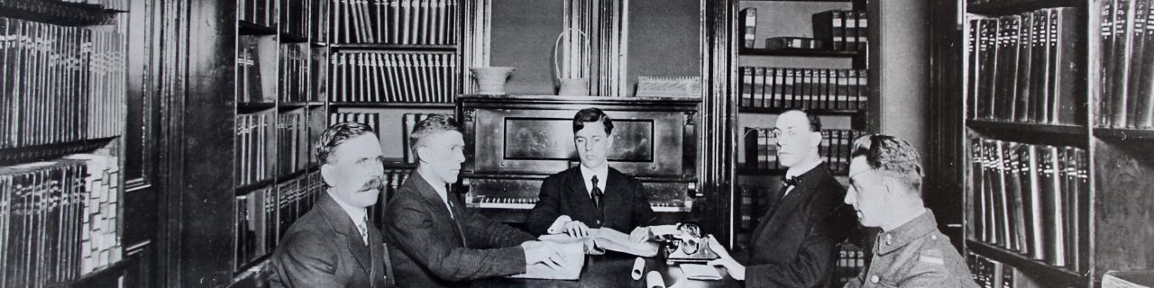 Five men, one in uniform, are seated around a table in the library with their hands on pages of braille. Dr. Carruthers has braille typewriter in front of him