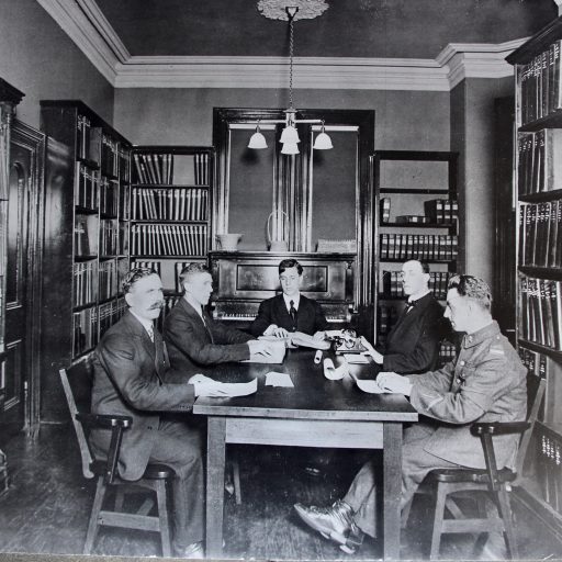 Five men, one in uniform, are seated around a table in the library with their hands on pages of braille. Dr. Carruthers has braille typewriter in front of him