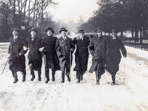 Seven men in a row with linked arms and carrying canes are running towards the camera on a snowy road. All the men are wearing hats and heavy greatcoats