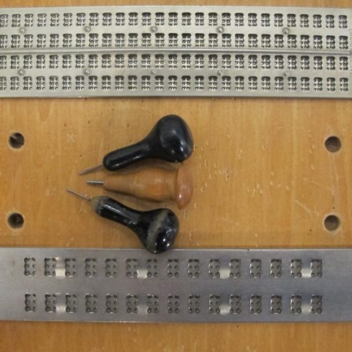 Upper slate supports four rows of 27 braille cells and the lower slate is for two rows of 15 cells. Three stylus examples lie on the wooden board which has guide holes at each side to stabilise the slate when in use