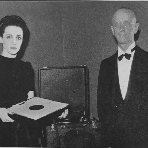 On the left Mlle. Curie is holding a box of phonograph disks, centre is an open phonograph machine, Dr. Swift stands on the right