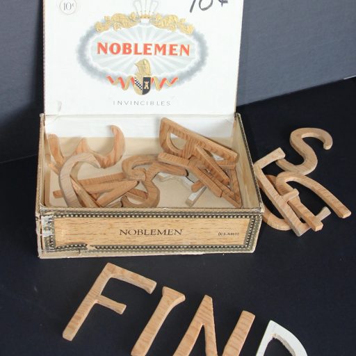 Une boîte à cigares ouverte contient des lettres en bois; il y a quelques lettres à côté de la boîte, et devant la boîte, le mot en lettres en bois FIND (trouver).