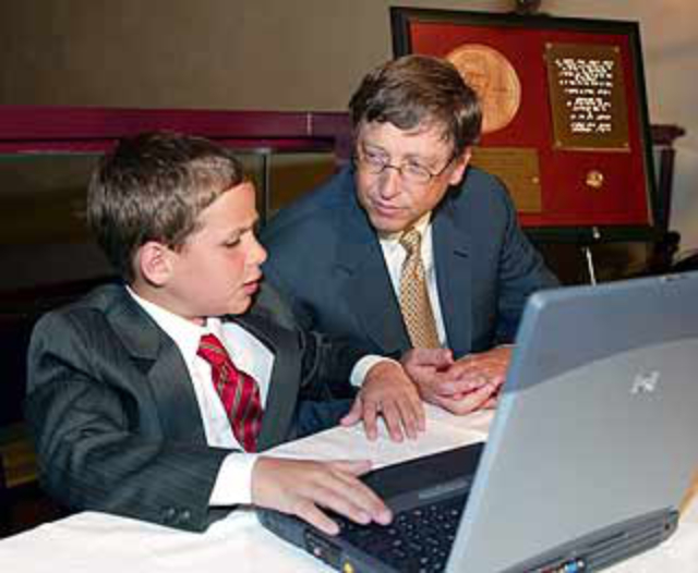Timothy has his hands on the keyboard of a laptop and Bill Gates is leaning over listening intently to Timothy