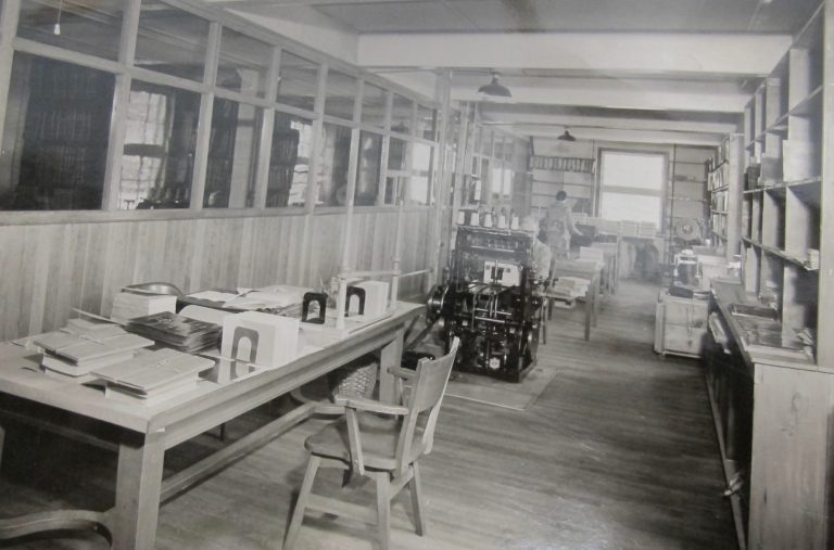 A long narrow room containing a table with finished products on it, a man sitting at a press and a woman with her back to the camera working at the end of the room