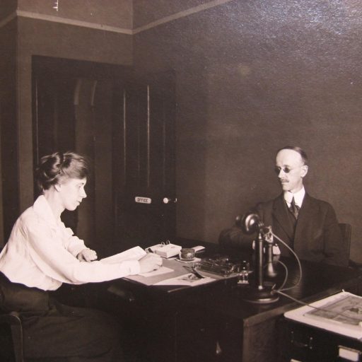 Son assistante écrit la dictée avec un stylo, M. Swift est assis de l’autre côté du bureau et un téléphone est entre eux sur le bureau.