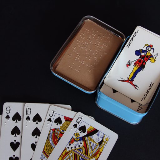 Braille playing cards - closeup of 6,9,10, jack and queen of spades with braille in top left corner, an open tin with braille instructions inside the lid and the joker on top of the pack