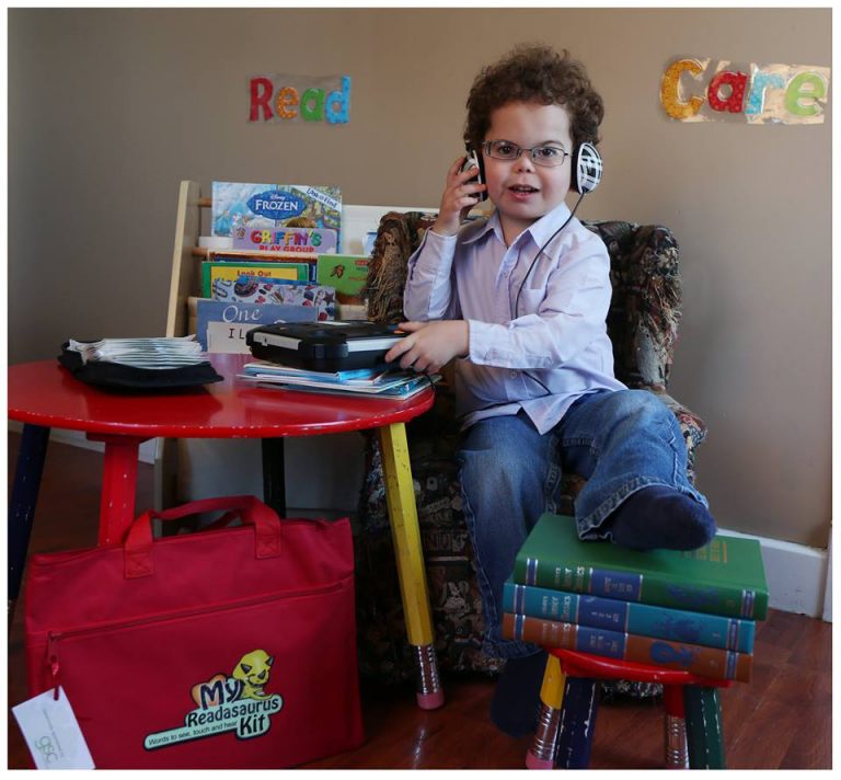 A young boy sits at a table with headphones on and a talking book player on the table. One foot is resing on a stool with several books on it and a red briefcase entitled My Readasauraus Kit is propped against the table leg. In the background tactile words - Read and Care - are on the wall and there is a rack of large print children's books